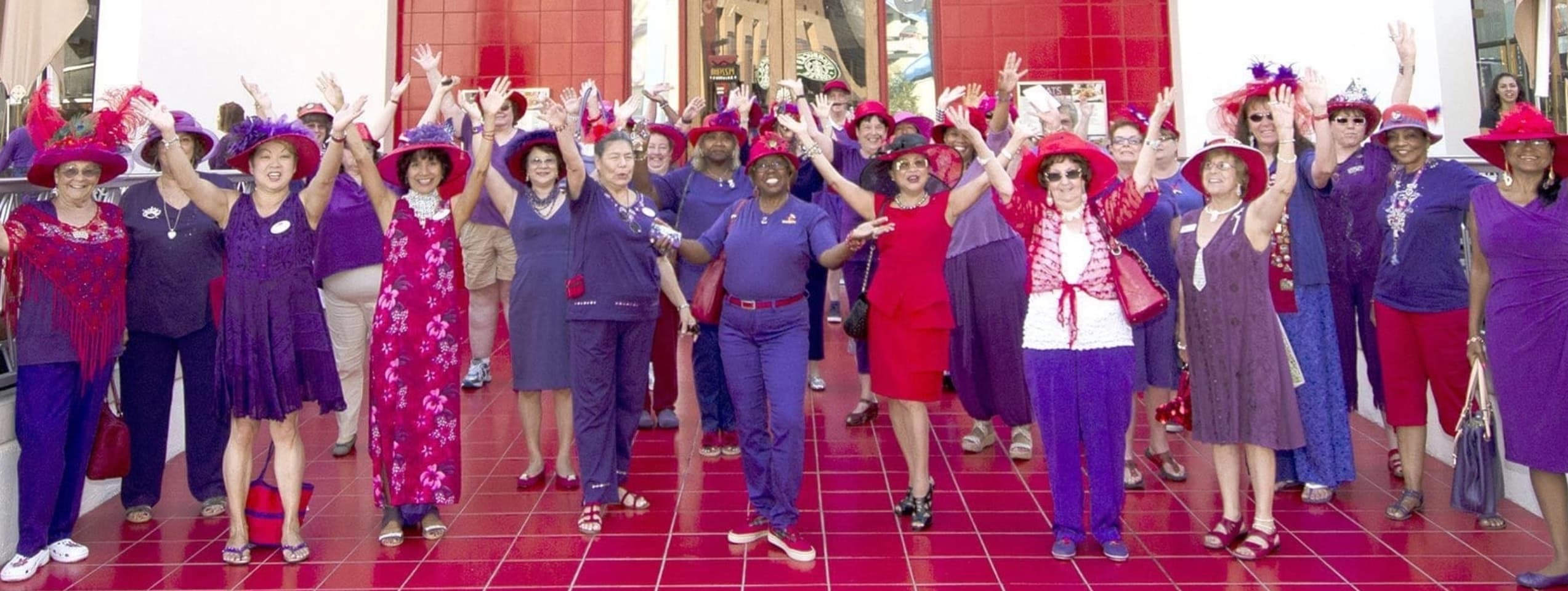 A large group of members in red hats and purple outfits cheering with their arms in the air on a red tiled plaza