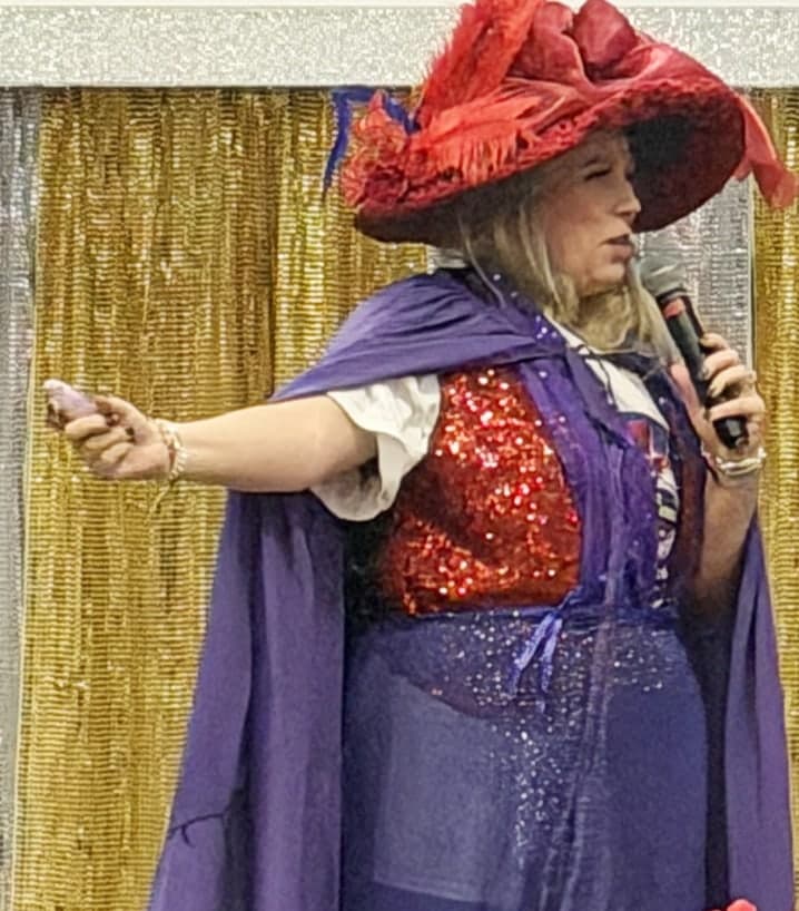Michelle Lee on stage in a wide red hat, a purple cape and a red sequined top, one arm outstretched