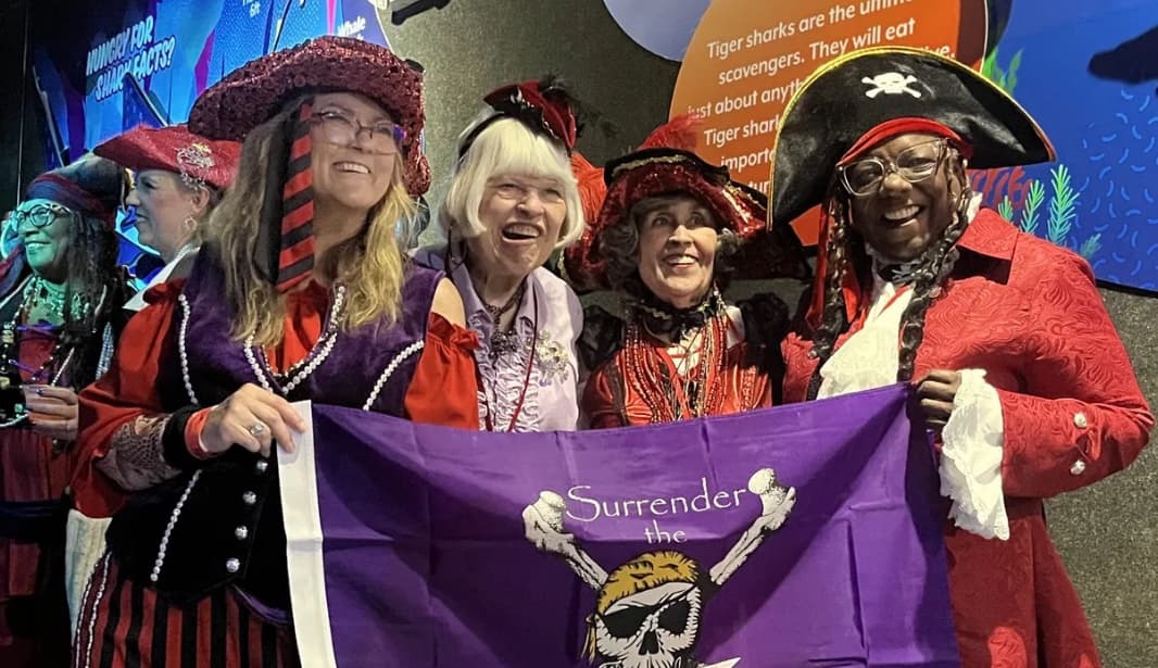 Five members in red hats and pirate costumes laughing together behind a purple Red Hat Society flag at a convention