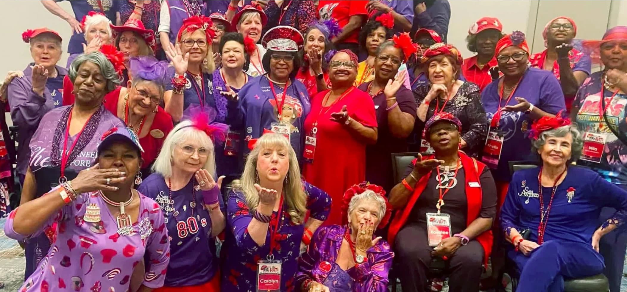 A large group of Red Hat Society members in red hats and purple outfits, gathered at a convention and blowing kisses to the camera.