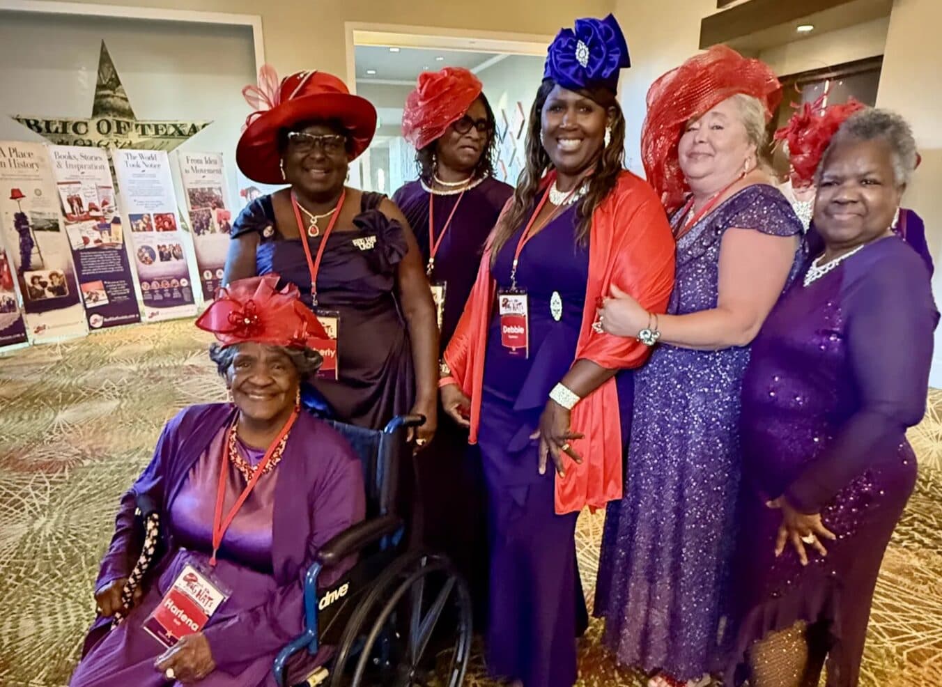 Six members in purple gowns and red hats posing together at a convention, one seated in a wheelchair at the front
