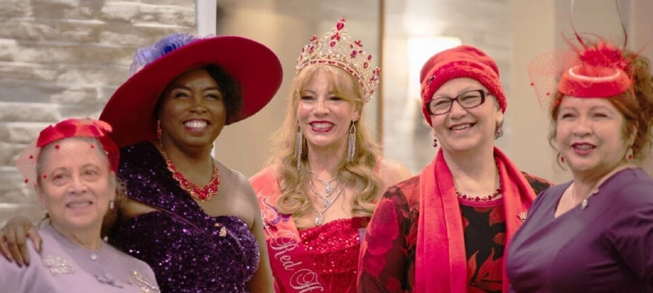 Five smiling members in red and purple, one wearing a jeweled crown and a sash, standing together at an indoor event